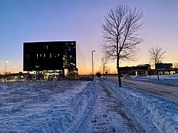 Black building facade with a sunset in the background; the sunset reflects off the side of the building. Snow covers the ground.