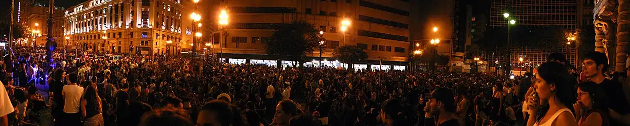 A huge crowd of people is gathered in a crowded public street at night in São Paulo outside the Municipal Theater
