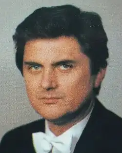 Colour publicity shot of a clean-shaven man with short, dark, curly hair, looking smoulderingly from under his eyebrows at the camera