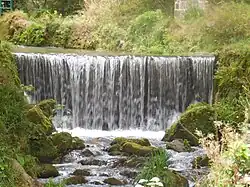 A weir with a river cutting a rough winding channel roughly 2 metres deep flowing through a wooded valley with paths.