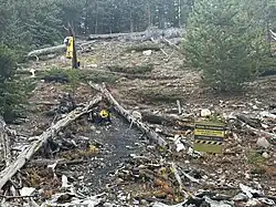 Pieces of airplane wreckage are scattered on the ground, along with a white cross and a yellow Wichita State football helmet. A sunflower and a small religious statue are visible as tributes. In the background, a sign reads, “Riding to Remember Wichita State University Football Team, October 2, 1970.” The area is rocky and covered with dry plants.