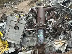 A large section of airplane wreckage lies scattered on a forested hillside. The debris consists of torn and twisted metal, with a piece of yellow fabric draped over part of it. More wreckage and fallen trees are visible in the background.