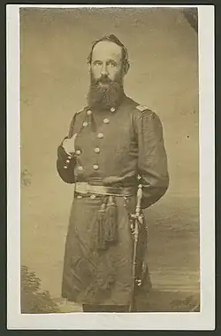 A standing bearded man in civil war uniform, sword at his side and right hand tucked in his jacket while left hand is behind his back. Posed for this photograph eyes looking at the camera and body slightly off square to the right from facing the camera.
