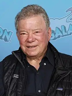Headshot of William Shatner in GalaxyCon Richmond. He is a later-aged, clean-shaven, light-skinned man with grey hair. He is wearing a button-up shirt and a jacket on top.