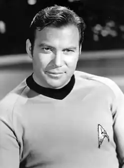 Catain James T. Kirk (played by Shatner) looks directly at the camera. He is wearing his uniform with the delta insignia.