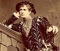 Terriss, in princely medieval costume with sword, leans against a stone bannister