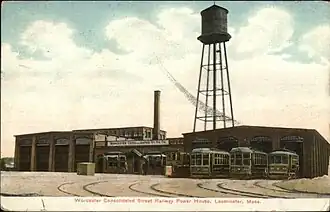 Postcard view of a brick streetcar barn with several streetcars visible. A water tower in the background is substantilaly taller than the building.