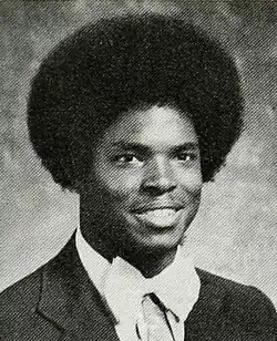 A Black teen is wearing a dinner jacket and white bow tie; with a large afro, he is facing right of the camera and smiling.
