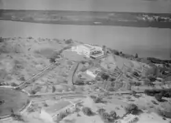 Black-and-white aerial photo of an area along a river with a few roads connecting a few buildings, with the largest building along the river.