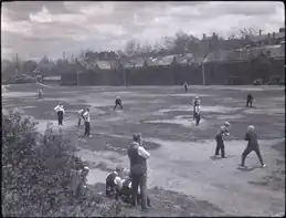 Men playing baseball. There are buildings, automobiles, and hydro lines in the background.
